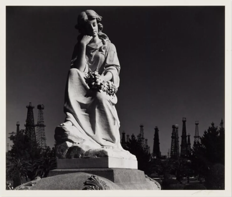 Photograph of a mourning female or angel statue atop a headstone with dark oil derrick silhouettes in the background.